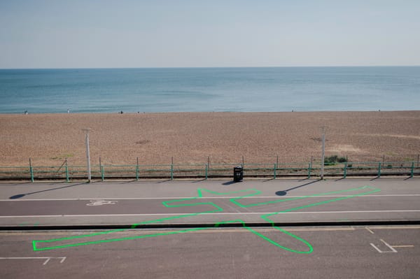 La sagome di un drone dipinta su un marciapiede di fronte a una spiaggia. Drone Shadow, un'opera di Jamse Bridle.